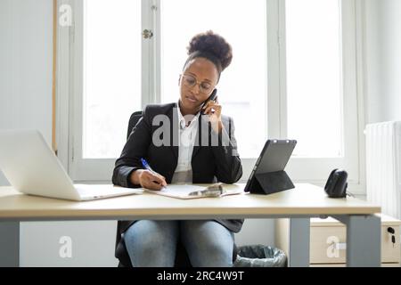 Concentrated young African American female bank manager speaking on phone and looking down while writing notes on clipboard and sitting at table with Stock Photo