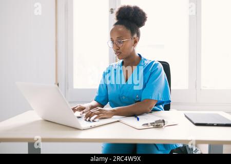 Focused young African American female doctor in blue uniform looking at screen of laptop and working while sitting at table with tablet and clipboard Stock Photo
