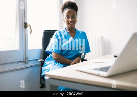 Positive African American female doctor in blue uniform and eyeglasses smiling and looking at camera while sitting at table with laptop in room with d Stock Photo