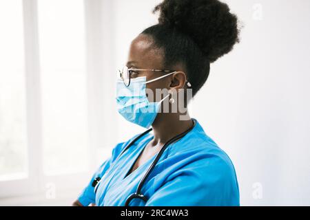 Side view of African American female physician in medical blue uniform and mask with stethoscope looking away thoughtfully in clinic Stock Photo