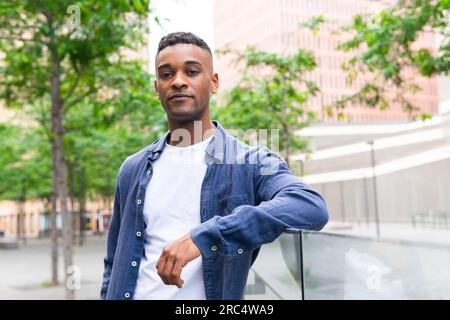 Portrait of self confident black male in casual clothes leaning on glass fence while chilling in park with green trees against cityscape and looking a Stock Photo