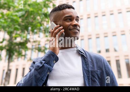 From below of smiling African American male entrepreneur in casual outfit smiling and talking on mobile phone while working on city street Stock Photo