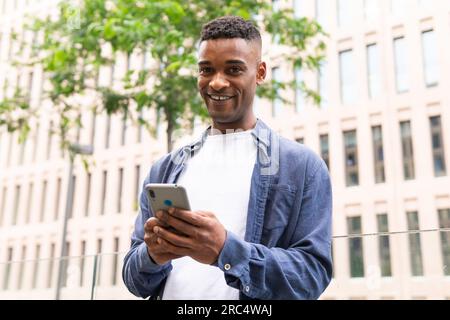 Smiling African American male entrepreneur in casual outfit smiling and text messaging on mobile phone while working on city street and looking at cam Stock Photo