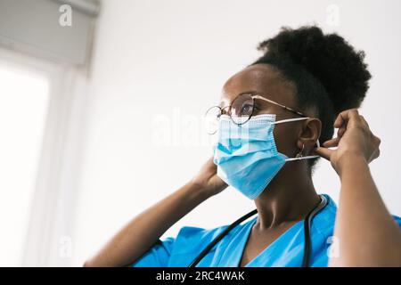 Side view of African American female physician in medical blue uniform with stethoscope looking away while putting on protective mask in hospital Stock Photo