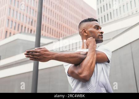 Low angle of serious African American male athlete in sportswear doing warming up exercises during workout and looking away in downtown of city Stock Photo