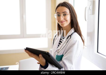 Young female doctor in white coat and eyeglasses looking at camera of tablet while standing in office of modern clinic Stock Photo