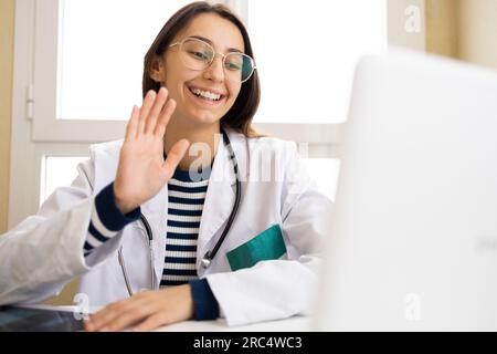 Positive young female doctor in white cloak waving hand to patient while having video call on laptop in office Stock Photo