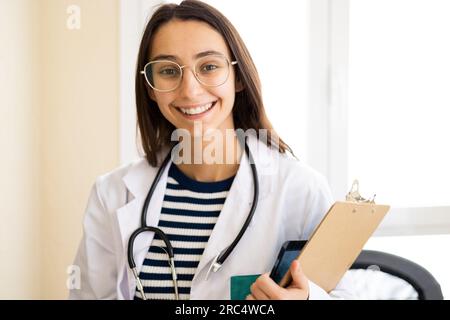 Positive young female doctor in white uniform and glasses smiling and looking at camera while holding clipboard Stock Photo
