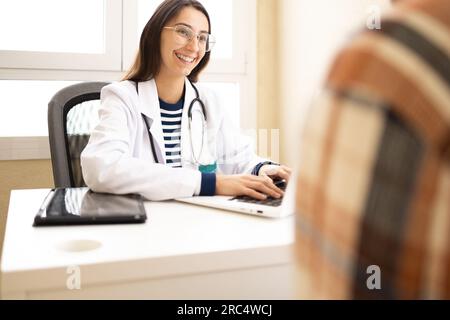 Cheerful young female doctor in white coat and glasses explaining treatment on paper document to crop patient while sitting at table Stock Photo