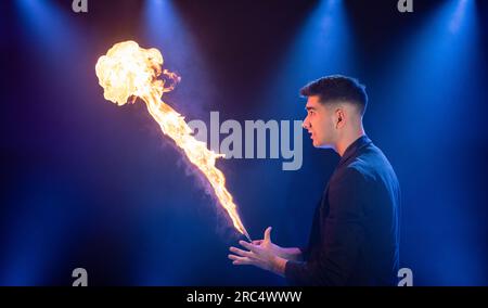 Side view of male prestidigitator in black suit showing magic trick with burning flame performing on dark stage in circus Stock Photo