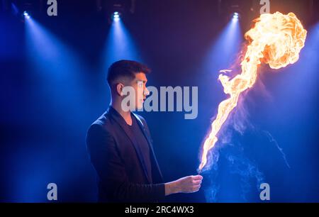 Side view of male prestidigitator in black suit showing magic trick with burning flame performing on dark stage in circus Stock Photo