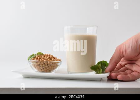 Detail of hand showing alternative soy drink on plate with full glass and bowl with dry beans on white table and white isolated background. Front view Stock Photo