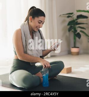 Smiling young female in sportswear with towel around neck using smartphone while sitting with bottle of energy drink on yoga mat at home Stock Photo