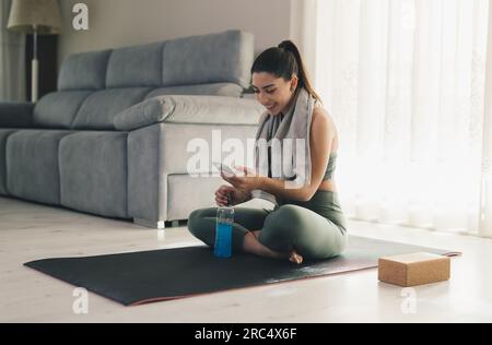 Smiling young female in sportswear with towel around neck using smartphone while sitting with bottle of energy drink on yoga mat at home Stock Photo