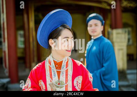 Portrait of positive young Asian female in traditional clothes and headwear looking away while standing against blurred background with man and buildi Stock Photo
