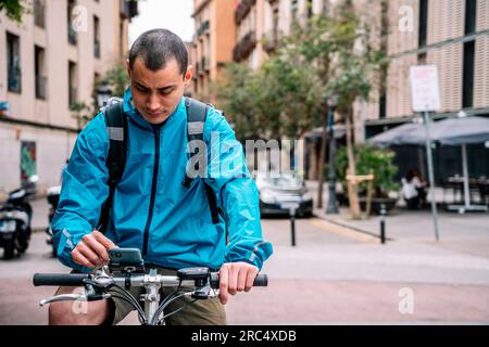 Delivery man in bright blue jacket with backpack sitting on bicycle in city street and using phone while working in Barcelona Stock Photo