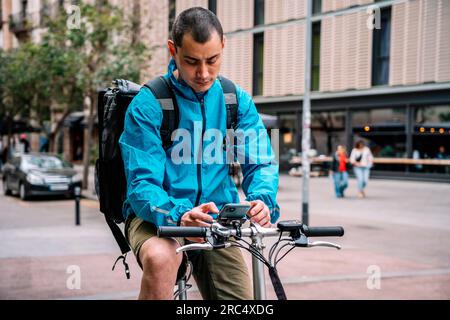 Delivery man in bright blue jacket with backpack sitting on bicycle in city street and using phone while working in Barcelona Stock Photo