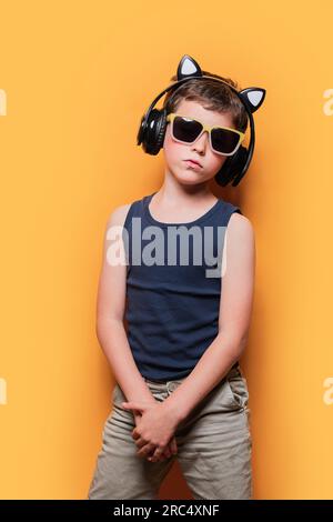 Confident boy in trendy outfit and sunglasses listening to music through wireless headphones against yellow background Stock Photo