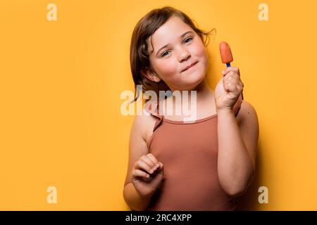 Adorable smiling girl in summer swimsuit holding delicious fruit ice pop against orange background while looking at camera Stock Photo