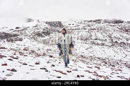 Full body of young bearded male traveler wearing hooded poncho looking away while walking on Chimborazo Volcano Ecuador against overcast sky during wi Stock Photo
