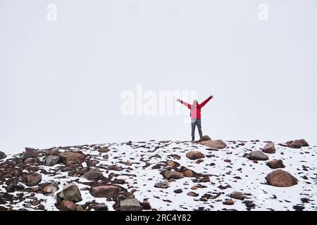 Full body of happy male in warm jacket with backpack with closed eyes while standing with outstretched hands on Chimborazo Volcano of Ecuador against Stock Photo