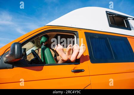 Crop anonymous people with legs stretched out through driver side window of yellow camper van against blue sky at daylight in canyon Rojo Teruel Spain Stock Photo