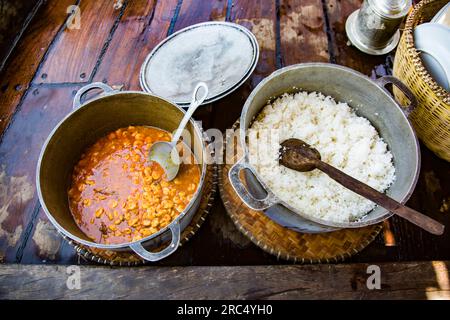 Top view of cooked rice and vegetable corn stew of Madagascar served in bowls with spoons and placed on wooden table with basket of ceramic plates Stock Photo