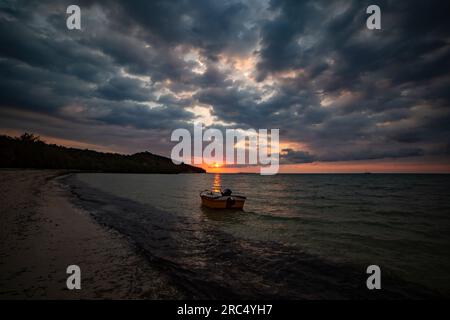 Silhouette of anonymous person in floating boat in seawater near sandy beach in Madagascar and enjoying orange sunset with black clouds in blue sky Stock Photo