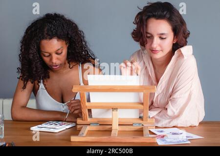 Concentrated young African American female sitting with diverse girlfriend at table with easel and looking down while using brushes and painting Stock Photo