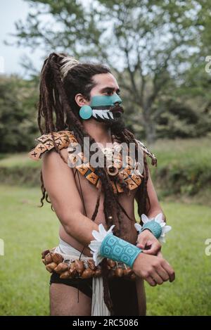 Confident braided male Mayan warrior in traditional costumes and body accessories with face painted standing on grass in park at daylight looking away Stock Photo