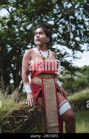 Low angle of young female Mayan warrior in stylish outfit with face and body painting looking away while standing in green park on sunny day Stock Photo