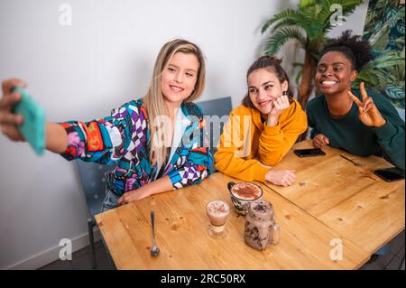 Group of cheerful multiracial friends in casual clothes sitting at table with cups of coffee and taking selfie on smartphone Stock Photo
