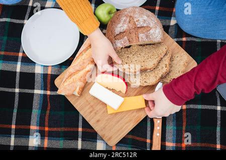 From above cropped unrecognizable women in casual clothes sitting on grass and eating bread with cheese while having picnic in autumn nature Stock Photo