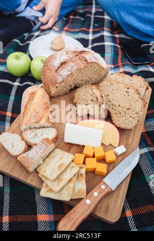 High angle of anonymous people sitting on plaid near knife and bread pieces with cut cheese on wooden board against apples Stock Photo