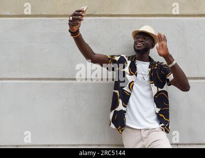 Stylish young African American male in colorful shirt and hat waving hand while taking selfie near stone wall in city Stock Photo