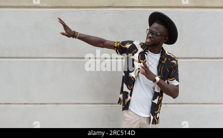 Delighted young African American guy in trendy outfit glasses and hat smiling and gesturing while standing against white wall on street Stock Photo