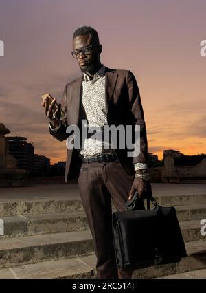 Concentrated young African American male entrepreneur in elegant stylish suit and eyeglasses with briefcase standing and using smartphone against suns Stock Photo