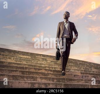 From below of full body of confident African American male in trendy formal suit and eyeglasses with briefcase walking on stairway in old town against Stock Photo