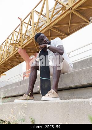 Low angle full body of pensive young African American male skater in casual clothes and sneakers leaning on skateboard and looking away thoughtfully w Stock Photo