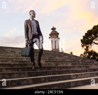 Low angle of self assured African American male in elegant suit and glasses carrying briefcase while standing on stairs and looking away Stock Photo