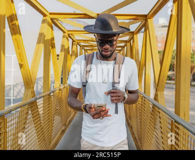Handsome modern African American guy in hat and with backpack surfing phone while strolling in city on yellow bridge Stock Photo