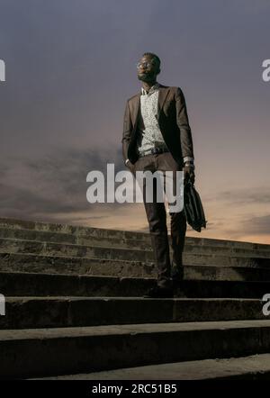 Low angle of self assured African American male in elegant suit and glasses carrying briefcase while standing on stairs and looking away with hand in Stock Photo