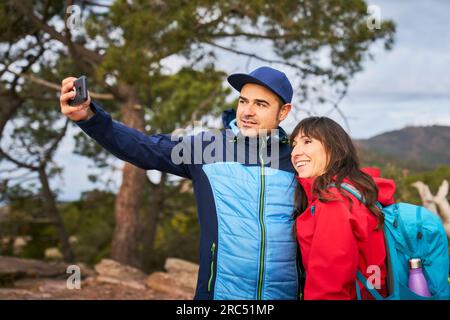 Glad wife and husband hugging and smiling while taking self portrait on smartphone with scenic view of El Garbi mountain Stock Photo