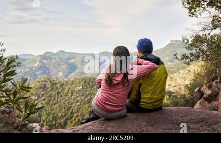 Back view of anonymous female traveler hugging boyfriend while sitting together on edge of cliff and admiring view of mountainous terrain Stock Photo