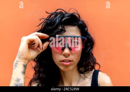Portrait of young Hispanic female touching red sunglasses with tattooed hand and looking at camera while in outside against orange wall in daylight Stock Photo
