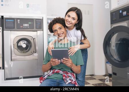 Couple Hugging Using Tablet At Laundrette While Washing Their Clothes Stock Photo