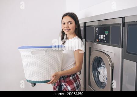 Happy Young Lady Doing Laundry At Laundrette, Posing Holding Basket Stock Photo