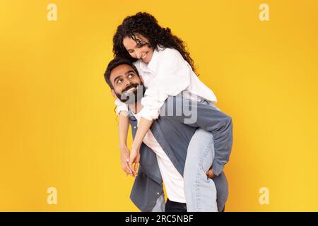 Cheerful Arabic Guy Carrying His Girlfriend Piggyback Over Yellow Background Stock Photo