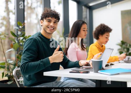 Group of multiracial students sitting at desk in classroom, selective focus on smiling european guy showing thumbs up Stock Photo