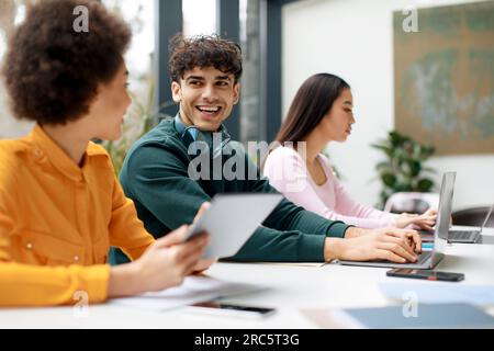 Happy diverse students sitting in classroom and communicating, using gadgets while being absorbed in studying Stock Photo